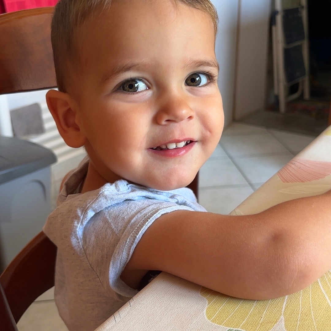 Léo participe au concours pour gagner de l'argent avec cette photo : arm, chair, child, clothing, door, eyes, face, furniture, hair, happy, indoor, kitchen, leaf, pattern, person, smiling, table, tablecloth, tile_floor, toddler