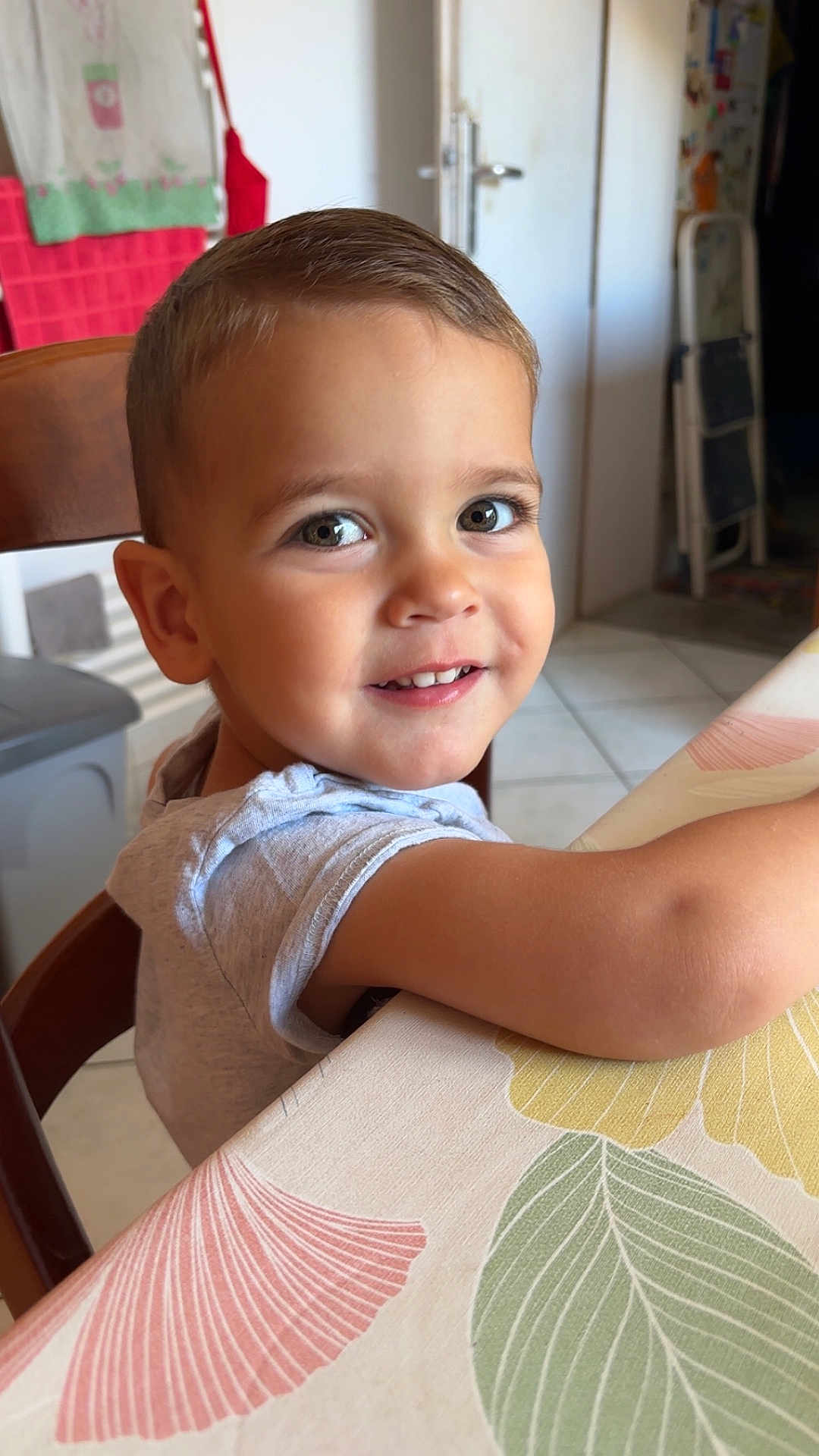 Léo participe au concours pour gagner de l'argent avec cette photo : child, toddler, smiling, face, eyes, table, chair, indoor, hair, clothing, arm, tablecloth, pattern, leaf, kitchen, tile_floor, door, furniture, person, happy