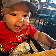 Jace joined the competition — help win amazing prizes! baby, child, cap, red_shirt, smiling, happy, indoor, restaurant, table, chair, blanket, person, face, cute, infant, clothing, wood, seat, laugh, headwear