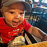baby, child, cap, red_shirt, smiling, happy, indoor, restaurant, table, chair, blanket, person, face, cute, infant, clothing, wood, seat, laugh, headwear