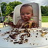 cake, child, chocolate, dirt, expressionless, face, food, greenery, hat, highchair, man, messy, nature, outdoor, person, seat, sky, summer, table, toddler