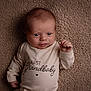 Easton is registered to the contest to win money with this photo: baby, infant, newborn, onesie, clothing, text, blanket, soft_texture, lying_down, portrait, cute, child, face, hand, expression, indoors, warm_colors, smiling, person, closeup