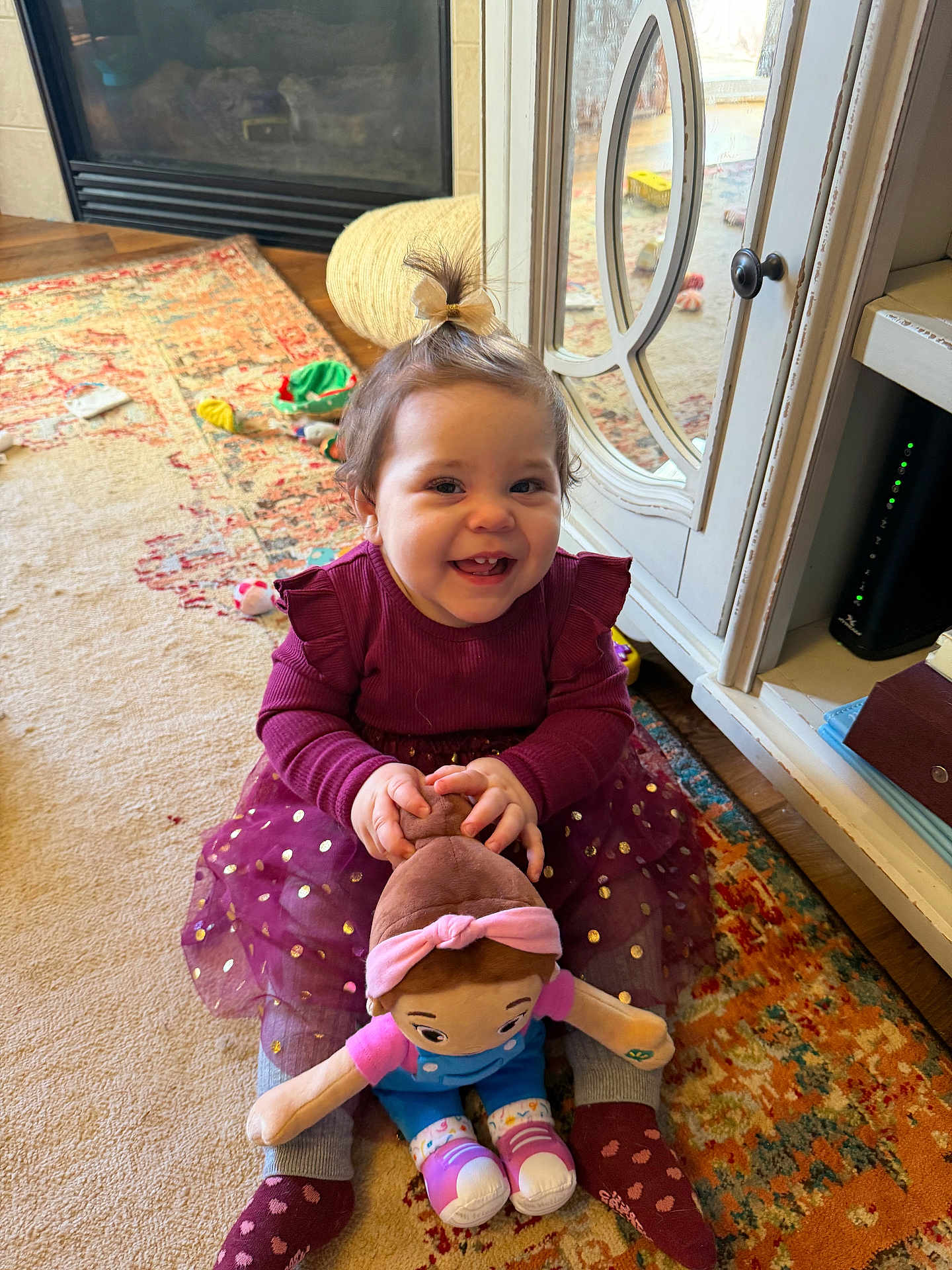 Adalyn is registered to the contest to win money with this photo: toddler, child, smiling, toy, doll, dress, hair_bow, socks, carpet, indoor, living_room, cabinet, wood_floor, happy, play, cute, person, furniture, colorful, cozy
