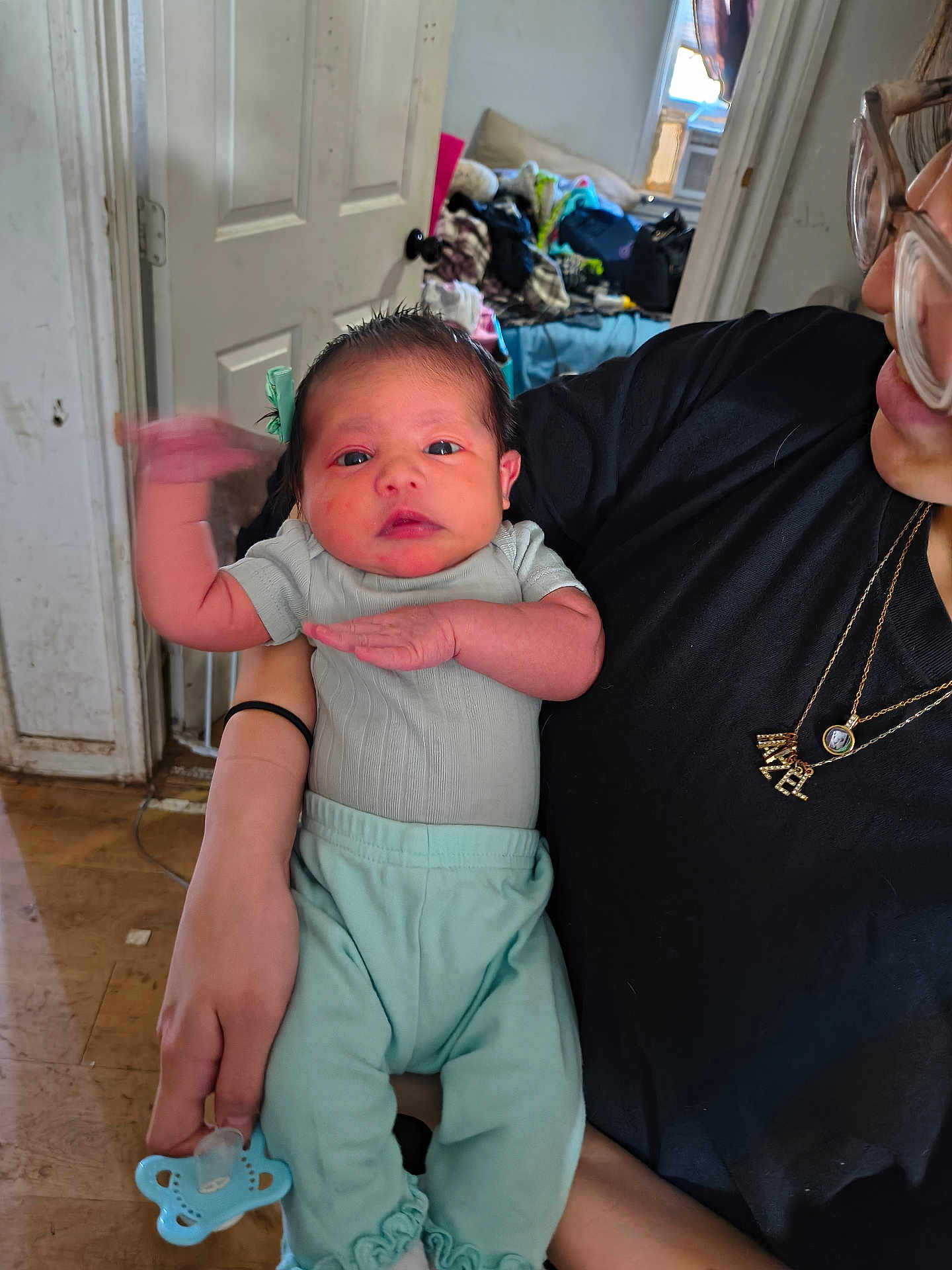 Heyzel is registered to the contest to win money with this photo: baby, infant, person, glasses, clothing, indoor, hand, face, necklace, door, room, pacifier, floor, window, furniture, black_shirt, mint_pants, holding, motion_blur, casual