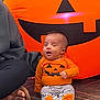 Desi is registered to the contest to win money with this photo: baby, infant, child, orange_clothing, striped_pants, pumpkin_face, halloween, inflatable_pumpkin, carpet, indoor, person, footwear, sitting, happy, cute, festive, celebration, smile, decor, adult