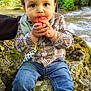 cap, child, curious, expression, face, food, fruit, greenery, hand, jeans, moss, nature, outdoor, plaid_shirt, rock, sitting, sneakers, stream, toddler, water