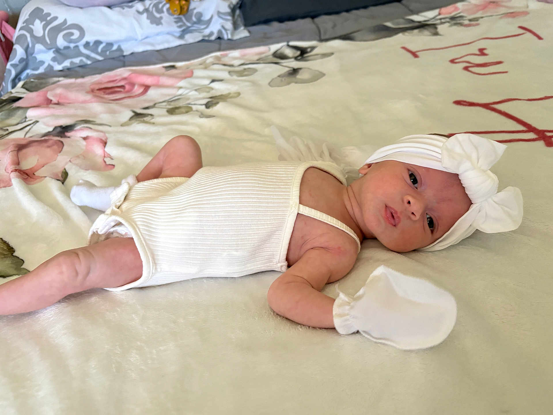 Irelynn is registered to the contest to win money with this photo: baby, infant, white_clothing, headband, bow, mittens, blanket, floral_pattern, lying_down, curious, newborn, face, human, soft_texture, bedroom, closeup, cute, small, resting, indoors