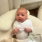 Raife is registered to the contest to win money with this photo: baby, infant, bib, cushion, plush_toy, star_teether, mirror, white_clothing, floor, curtain, radiator, indoor, child, cute, soft_texture, hand, face, expression, newborn, comfort