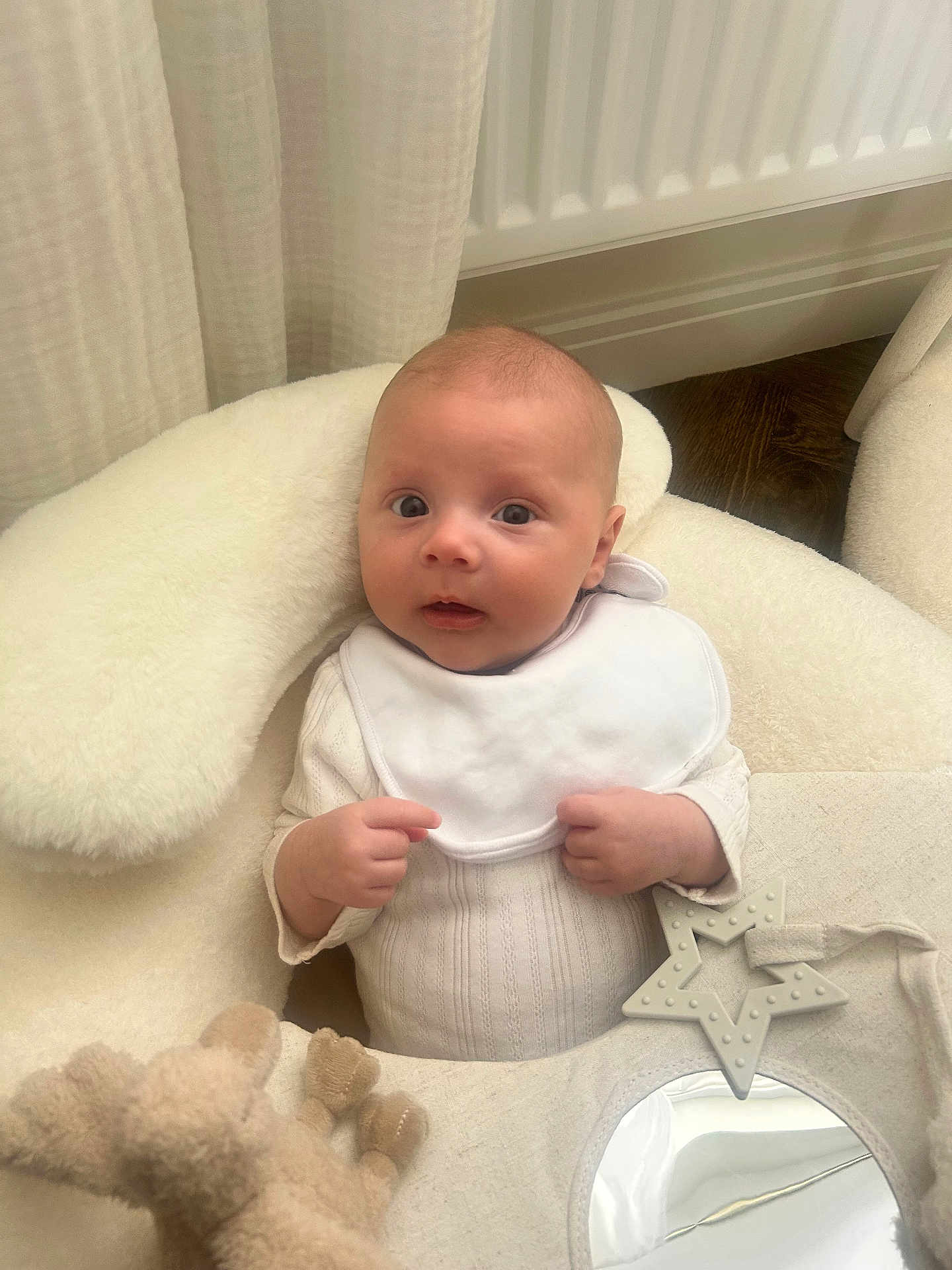 Raife is registered to the contest to win money with this photo: baby, infant, bib, cushion, plush_toy, star_teether, mirror, white_clothing, floor, curtain, radiator, indoor, child, cute, soft_texture, hand, face, expression, newborn, comfort
