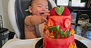 Armando is registered to the contest to win money with this photo: baby, blinds, celebration, child, clothing, food, fruit_cake, happy, high_chair, home, indoor, orange_slices, person, plate, smiling, strawberries, table, watermelon, window, wood_floor