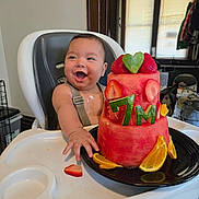 Armando joined the competition — help win amazing prizes! baby, high_chair, watermelon_cake, strawberries, orange_slices, fruit_cake, happy, smiling, indoor, milestone, celebration, child, person, food, plate, window, clothing, hand, face, home