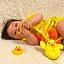 baby, child, yellow_clothing, toy, rubber_duck, plush_toy, blanket, cute, infant, lying_down, soft_texture, indoors, playtime, smiling, face, hand, bright_color, childhood, adorable, young