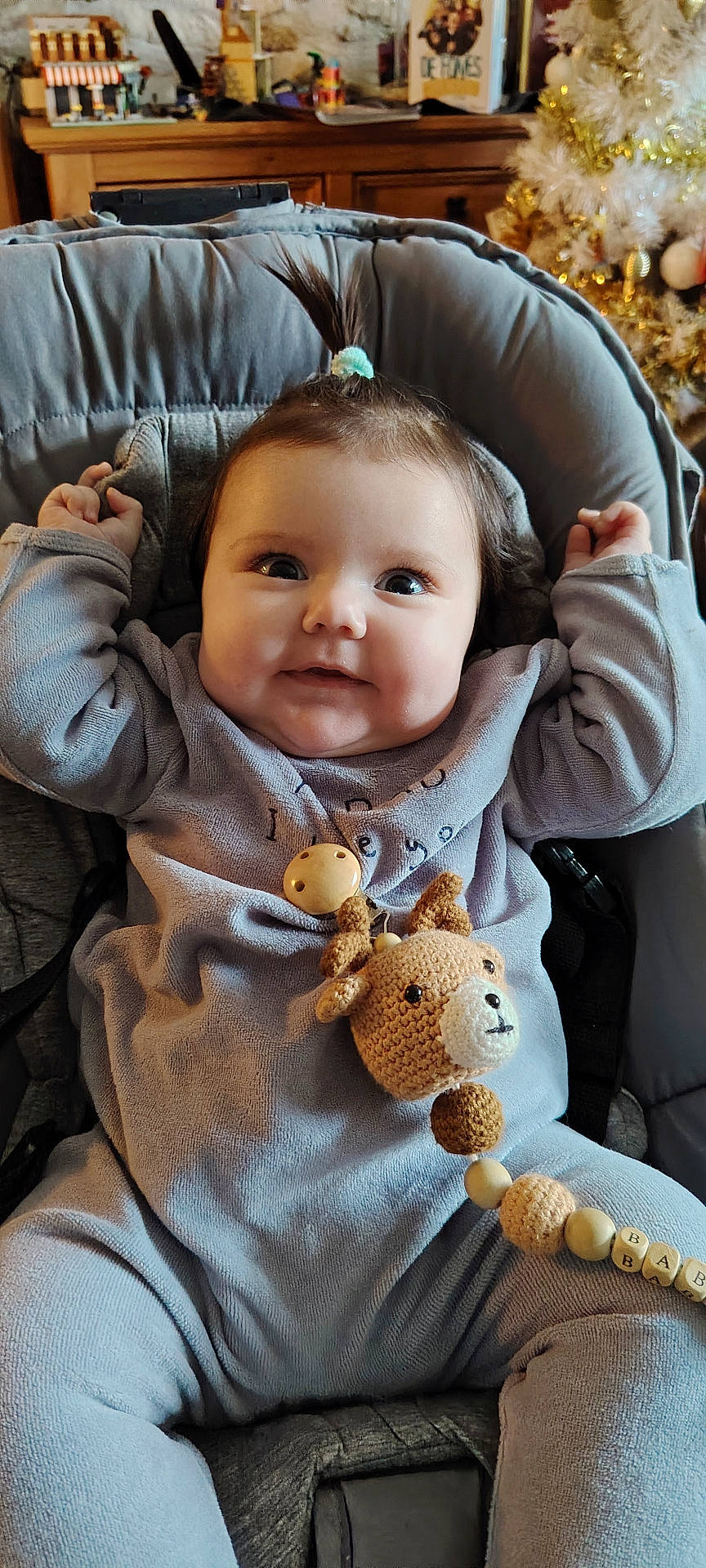 baby, baby_seat, beads, child, christmas_tree, crocheted_toy, cushion, cute, face, festive, happy, indoor, infant, ornaments, pony_tail, purple_outfit, reindeer_toy, smiling, warm_lighting, wooden_furniture