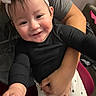adult_hand, arm, baby, black_shirt, child, closeup, clothing, cute, face, happy, headband, holding, human, indoor, person, portrait, reaching_hand, sitting, smile, white_pants