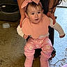 baby, infant, pink_bow, pink_bib, pink_boots, floral_pants, curious_expression, hands_supporting, indoor, concrete_floor, adult_arms, long_hair, child, clothing, footwear, person, floor, cute, standing, support