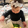 baby, black_shirt, blanket, book, bow, carpet, child, cozy, cute, face, hand, headband, indoor, infant, person, plush_toy, reading, sitting, soft_lighting, toy