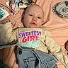 baby, infant, clothing, denim, bed, fabric, face, person, text, shirt, lying_down, bedspread, diaper, colorful_text, casual, indoors, cute, skin, expression, newborn