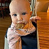 baby, child, cookie, blue_eyes, pink_clothing, unicorn_pattern, highchair, wooden_table, indoor, restaurant, face, hand, curious, food, messy, person, portrait, seat, table, cute
