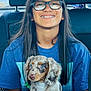 person, child, glasses, braces, puppy, dog, car_interior, seat, blue_shirt, long_hair, smile, young, pet, holding, cute, sitting, window, daylight, happy, animal
