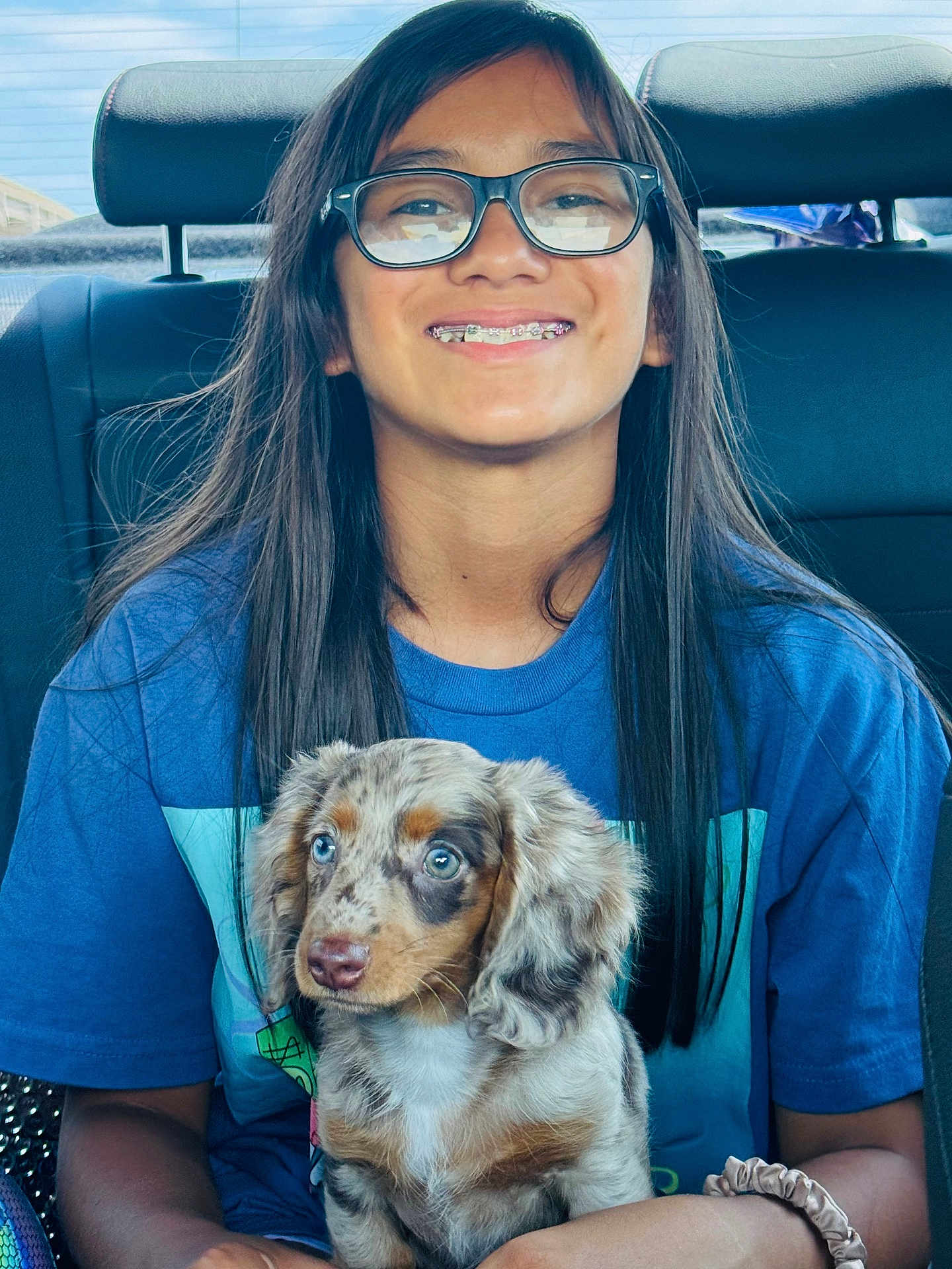 Piper Morones joined the competition — help win amazing prizes! person, child, glasses, braces, puppy, dog, car_interior, seat, blue_shirt, long_hair, smile, young, pet, holding, cute, sitting, window, daylight, happy, animal