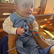 Marli Lima Tedesco participe au concours pour gagner de l'argent avec cette photo : baby, child, infant, blue_suit, striped_clothing, hands, tattoo, bracelet, church, wooden_pews, sunlight, indoor, toy, curious, holding, person, face, eyes, clothing, seated