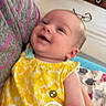 Madelyn is registered to the contest to win money with this photo: baby, child, yellow_dress, smiling, indoor, bedroom, furniture, person, happy, face, holding, wooden_furniture, blanket, glasses, social_media, mobile_screen, text_overlay, portrait, infant, casual