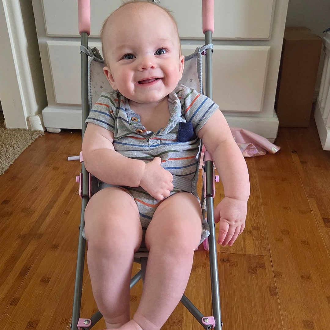 Lucas is registered to the contest to win money with this photo: baby, smiling, stroller, indoor, wooden_floor, dresser, child, onesie, pink, person, happy, infant, furniture, floor, legs, feet, face, hand, head, toy