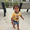 child, toddler, boy, curly_hair, barefoot, standing, outdoor, boardwalk, beach, dune_grass, bench, yellow_shirt, cowboy_shirt, shorts, summer, portrait, person, face, playful, daylight