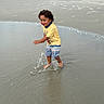 Armani is registered to the contest to win money with this photo: child, toddler, beach, ocean, seashore, water, splashing, wet_sand, outdoor, smiling, curly_hair, yellow_shirt, shorts, barefoot, playing, summer, waves, shoreline, beachgoer, portrait