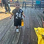 child, toddler, football_jersey, face_paint, outdoor, wooden_deck, adult, sports_fan, casual_clothing, daylight, chairs, table, string_lights, smiling, person, walking, yellow, black, shoes, hat