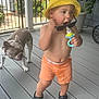 Lorenzo is registered to the contest to win money with this photo: toddler, child, yellow_hat, orange_shorts, pacifier, sunglasses, dog, pet, porch, deck, wooden_floor, summer, bare_chest, footwear, black_clogs, plant, potted_plant, fence, outdoor, daylight