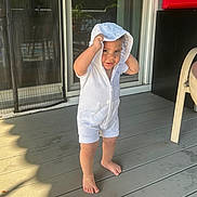 Lorenzo is registered to the contest to win money with this photo: toddler, child, hoodie, white_clothing, barefoot, porch, deck, glass_table, chair, sunlight, outdoor, curious, playful, smiling, person, young_child, casual, daylight, standing, expression
