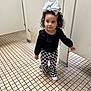 Oaklyn is registered to the contest to win money with this photo: toddler, child, curly_hair, bow, black_top, checkered_pants, white_shoes, bathroom, tiles, floor, stall, door, person, indoor, cute, fashion, walking, exploring, young_child, curiosity