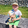 bench, blue_sneakers, boy, cap, casual_clothing, child, clouds, grass, green_shorts, happy, monkey, nature, outdoor, playful, portrait, sky, stone_wall, stuffed_toy, summer, sunny_day