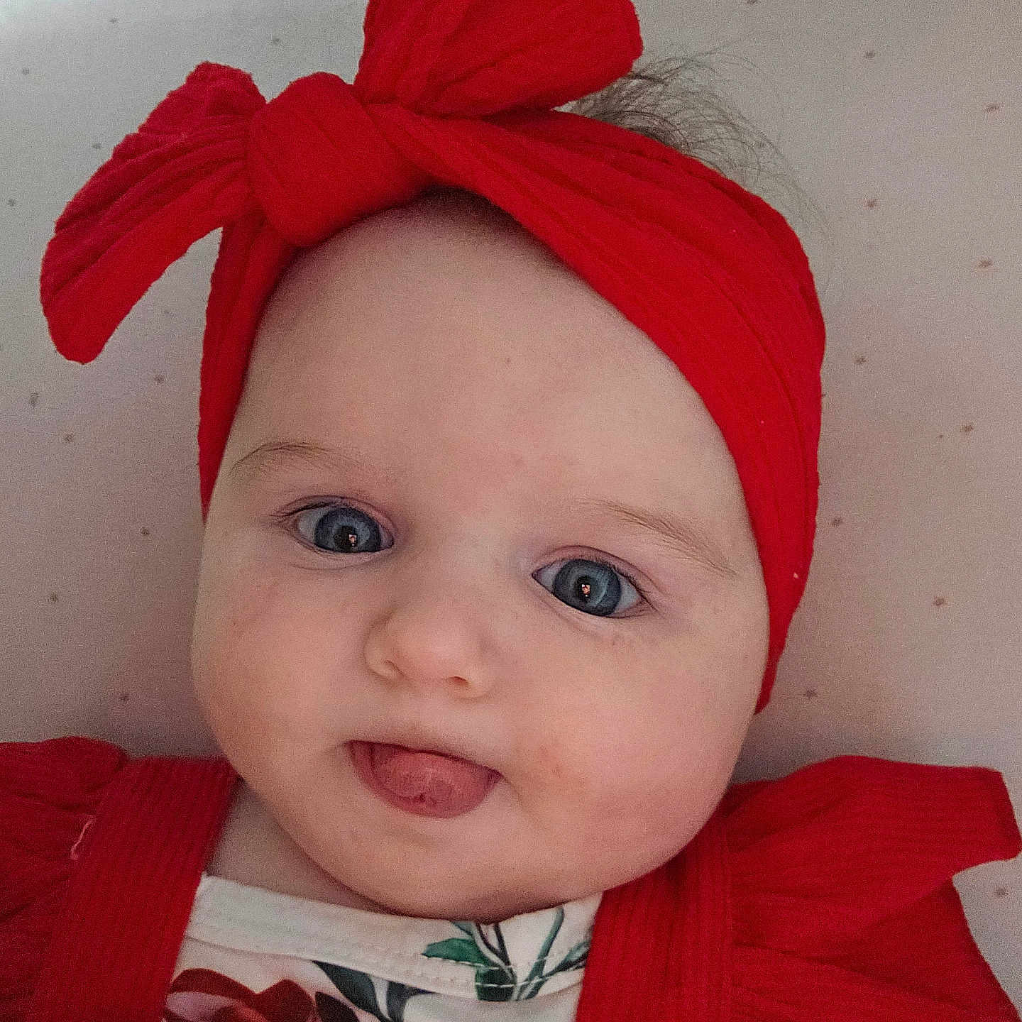Inaë a rejoint le concours — aidez-le/la à gagner de superbes lots ! baby, child, face, headband, red_clothing, tongue_out, blue_eyes, cute, infant, portrait, lying_down, closeup, soft_lighting, playful, smiling, indoors, floral_pattern, headwear, red_bow, cheeks