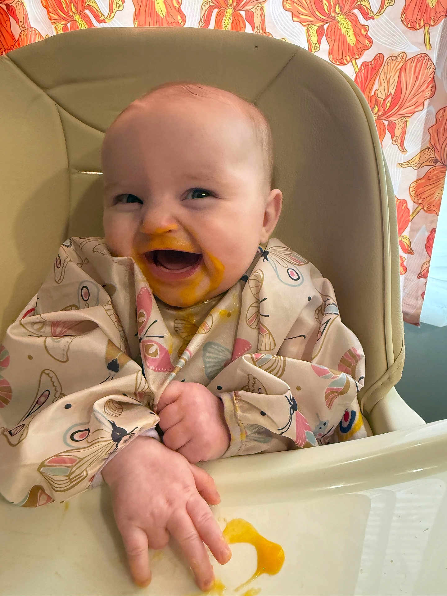 Lydia is registered to the contest to win money with this photo: baby, high_chair, food, messy, bib, butterfly_pattern, smiling, happy, hands, orange_food, infant, indoor, child, cute, feeding, playful, face, clothing, expression, young