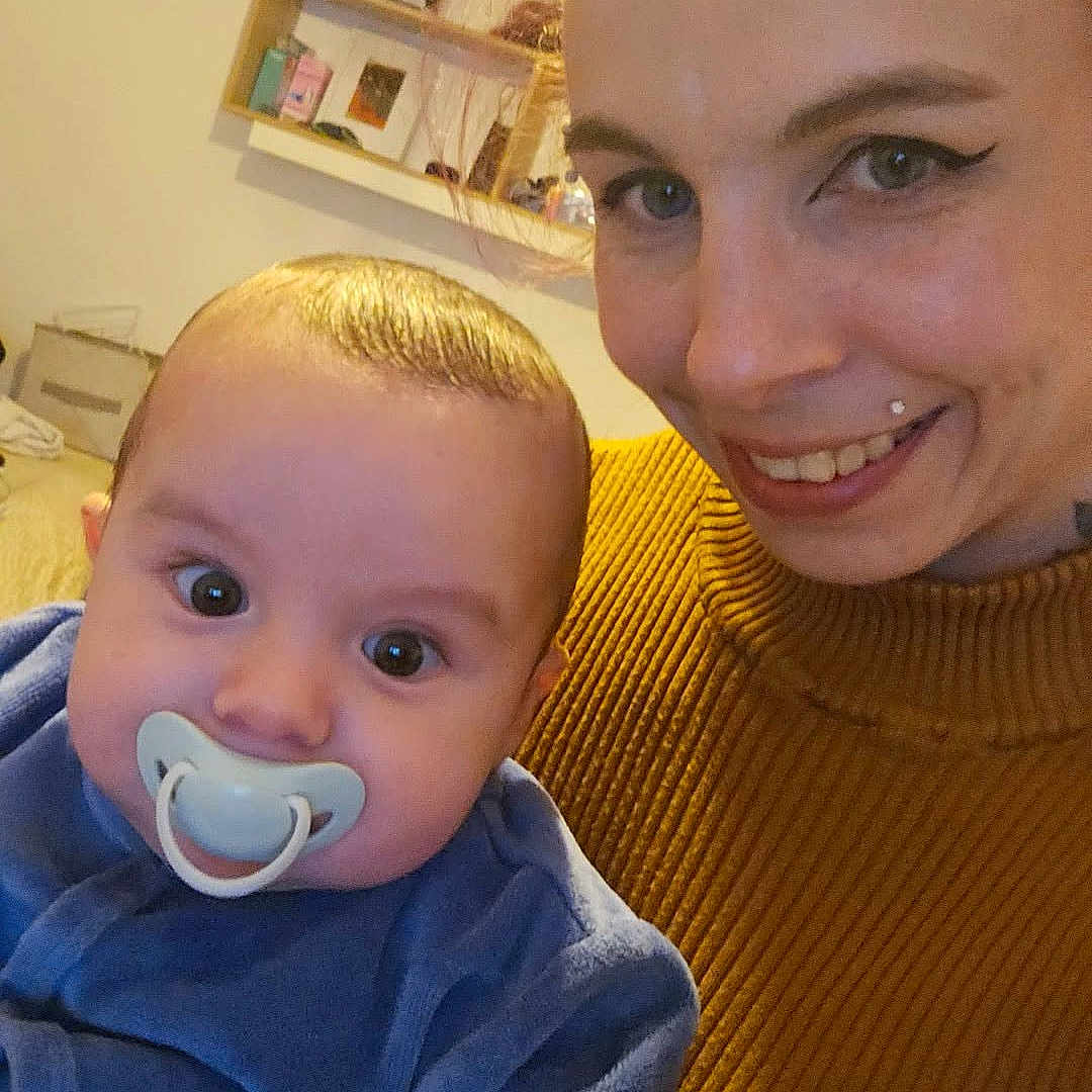 Thyago Rambert a rejoint le concours — aidez-le/la à gagner de superbes lots ! adult, baby, close_up, cozy, eyes, face, hair, holding, indoor, infant, living_room, pacifier, picture_frame, portrait, shelf, skin, smile, striped_sweater, sweater, woman