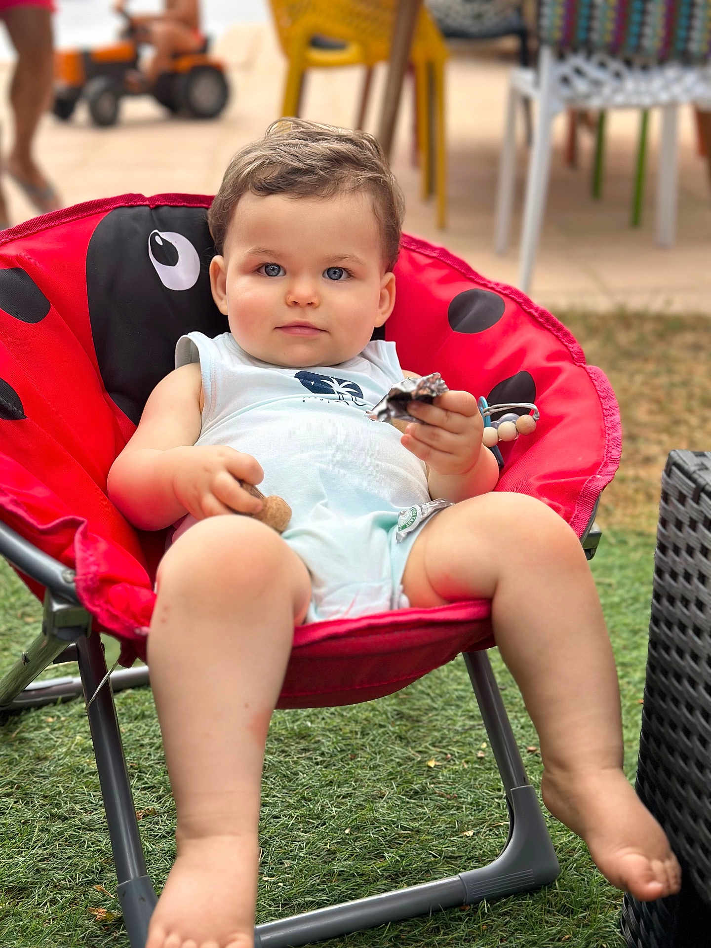 Andrea a rejoint le concours — aidez-le/la à gagner de superbes lots ! toddler, child, baby, red_chair, ladybug_pattern, outdoor, grass, snack, toy, blue_eyes, curly_hair, summer_clothing, barefoot, plastic_chair, relaxed, portrait, seat, playtime, casual, sunlight