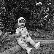 Elizabeth is registered to the contest to win money with this photo: toddler, child, overalls, headband, shoes, garden, bushes, flower, grass, wood, outdoor, smile, sitting, nature, portrait, young_child, person, plants, black_and_white, casual_clothing