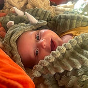 Teo joined the competition — help win amazing prizes! baby, child, costume, dinosaur, fuzzy, blanket, smile, face, head, happy, infant, cute, warm, cozy, soft, pillow, indoor, portrait, clothing, expression