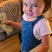 Paisley joined the competition — help win amazing prizes! toddler, child, money, cash, smile, denim_overalls, pink_shirt, indoor, kitchen_counter, wood_floor, plant, television, furniture, happy, portrait, hand, face, person, home, cute