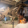 toddler, child, baby, person, face, portrait, museum, dinosaur, skeleton, fossil, exhibit, indoors, orange_dress, floral_pattern, adult, parent, arms, zoo, educational, photography