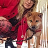 woman, dog, shiba_inu, indoor, leash, glasses, red_jacket, plaid_pants, wooden_fence, floor, footwear, black_boots, necklace, pet, crouching, light, fence, animal, person, casual