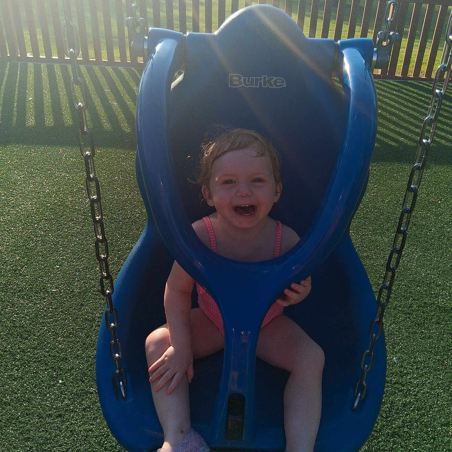 Riley is registered to the contest to win money with this photo: toddler, child, swing, playground, outdoor, smiling, joyful, blue, sunlight, grass, fence, park, summer, happy, seat, plastic, play, fun, person, casual