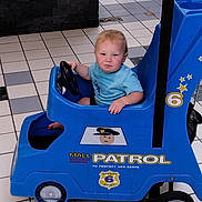 Jaxon is registered to the contest to win money with this photo: toddler, child, toy_car, mall, indoor, blue, steering_wheel, patrol, serious_expression, floor_tiles, short_sleeve_shirt, blonde_hair, foot, plastic, wheels, badge, number_6, safety, person, play