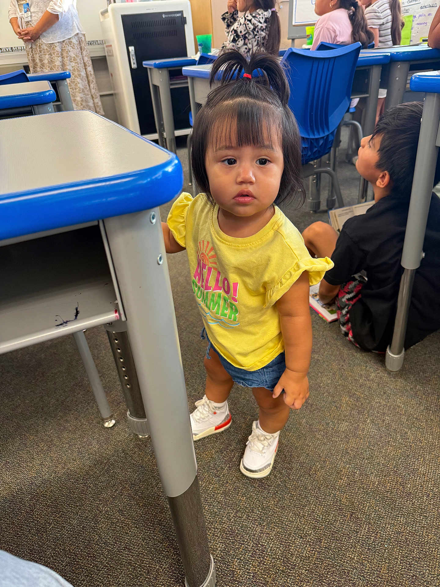 Mia is registered to the contest to win money with this photo: child, toddler, classroom, desk, yellow_shirt, white_sneakers, curious, standing, children, teacher, carpet, blue_chair, hair_tied, shorts, indoor, school, person, learning, education, young