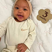 Zaniyah is registered to the contest to win money with this photo: baby, infant, smiling, headband, bow, beige_clothing, onesie, wooden_sign, 3_months, blanket, pink_bows, lying_down, cute, happy, portrait, closeup, child, person, indoors, soft_lighting