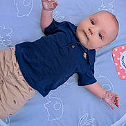 Kai is registered to the contest to win money with this photo: baby, infant, blue_shirt, beige_shorts, milestone_card, 3_months, blanket, pattern, lying_down, cute, child, person, indoor, soft_light, calm, curious, small_hands, short_hair, face, eyes