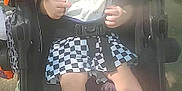 Kai is registered to the contest to win money with this photo: baby, child, stroller, sneakers, checkered_skirt, bib, sunlight, outdoor, happy, smiling, person, footwear, seat_belt, infant, clothing, face, hands, legs, toy, bottle