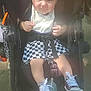 baby, child, stroller, sneakers, checkered_skirt, bib, sunlight, outdoor, happy, smiling, person, footwear, seat_belt, infant, clothing, face, hands, legs, toy, bottle